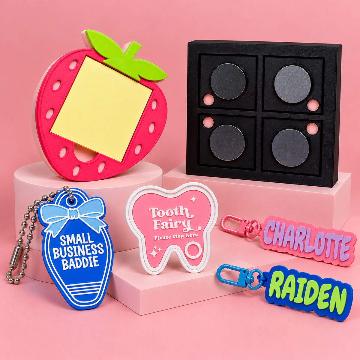 3D-printed gifts and accessories by Printed To Stick, featuring a strawberry memo holder, black magnetic organiser, Tooth Fairy tray, Small Business Baddie keyring and personalised name keyrings displayed on pink podiums.