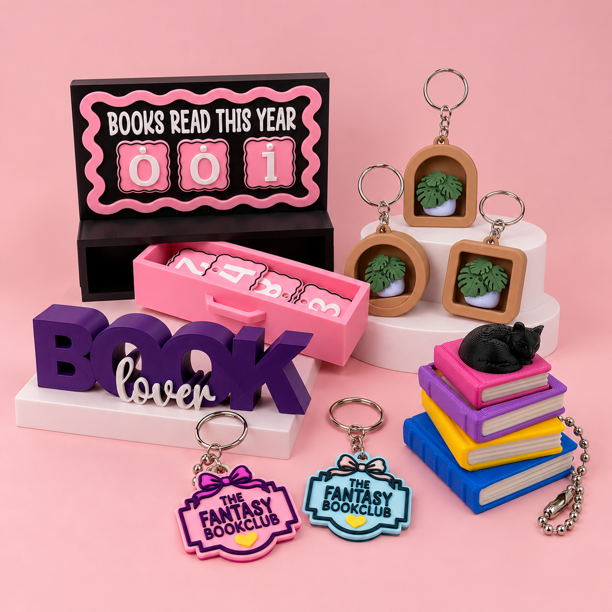 3D printed bookish gifts including a books read tracker, book lover sign, miniature book stack with black cat, monstera plant keyrings and Fantasy Bookclub keyrings in pink, purple and blue.